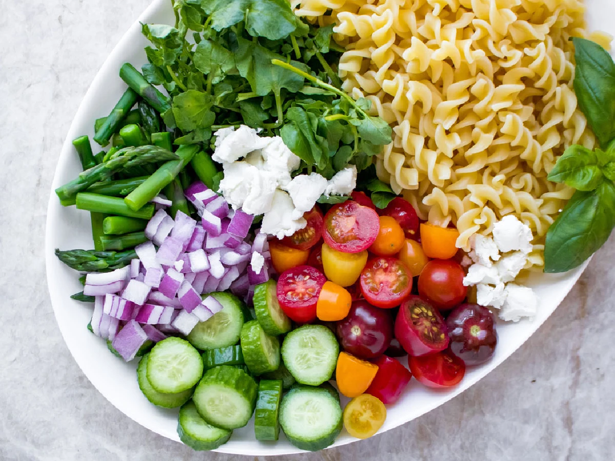 A platter with uncooked pasta, cherry tomatoes, cucumber slices, diced red onion, asparagus, leafy greens, basil, and dollops of white cheese.