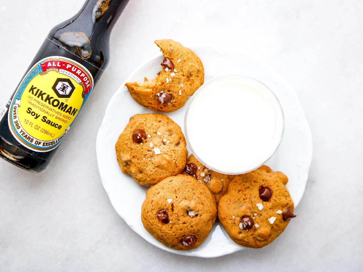A bottle of Kikkoman soy sauce next to a plate of chocolate chip cookies and a glass of milk on a white surface.