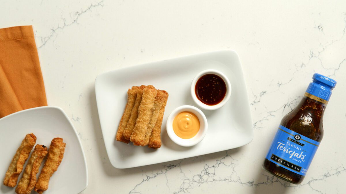 Rectangular plate with four fried spring rolls, small bowls of yellow and brown sauces, a bottle of teriyaki marinade, and an orange napkin on a white marble surface.