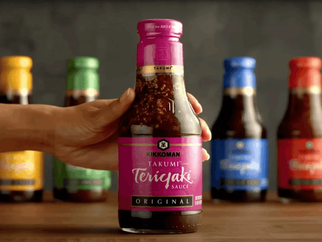 A hand holds a bottle of Kikkoman Takumi Teriyaki Sauce Original flavor, with other sauce bottles blurred in the background.