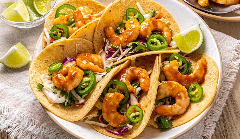 Four shrimp tacos topped with sliced jalapeños, cabbage slaw, and sauce are arranged on a white plate with lime wedges on the side.