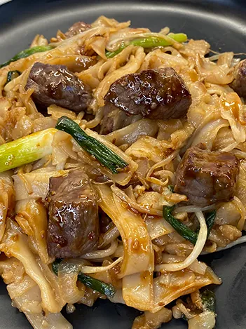 A plate of stir-fried flat rice noodles with beef, green onions, and bean sprouts in a savory sauce.