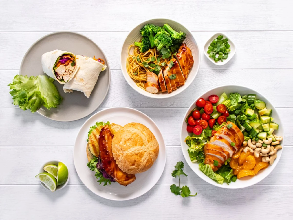 A table with four plates of food, including a wrap, chicken salad, noodles with chicken and broccoli, and a biscuit with glazed chicken, plus a small bowl of green onions and lime wedges.