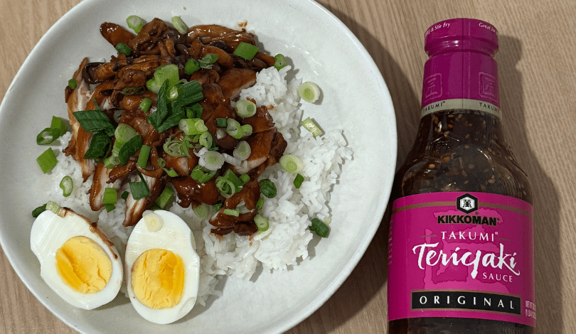 A plate of white rice topped with sliced chicken, chopped green onions, and a halved boiled egg, beside a bottle of Kikkoman Takumi Teriyaki Sauce.