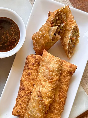 Three crispy egg rolls on a rectangular white plate, one cut open to show the filling, served with a small bowl of dipping sauce.