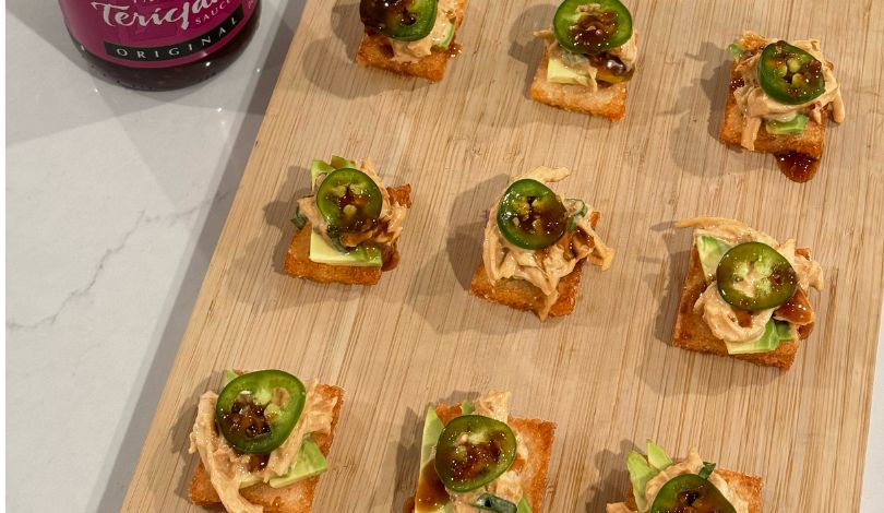 Nine square appetizers topped with avocado, shredded chicken, sliced jalapeño, and sauce are arranged on a wooden board, with a bottle of teriyaki sauce visible in the corner.