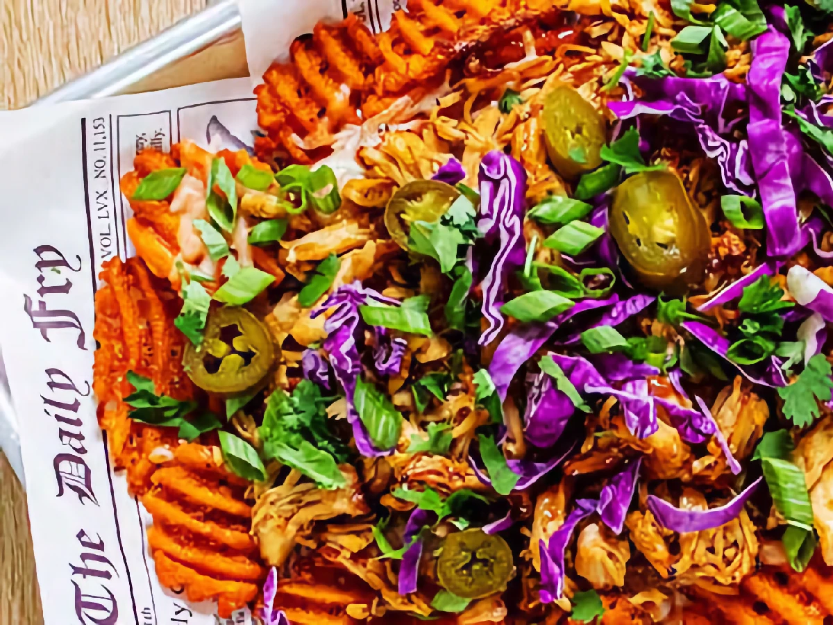 A serving of waffle fries topped with shredded chicken, purple cabbage, jalapeños, green onions, and cilantro on newspaper-style parchment paper.