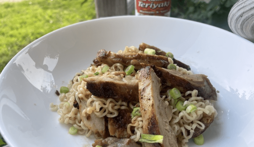 A white bowl containing ramen noodles topped with sliced grilled chicken and chopped green onions, with a bottle of teriyaki sauce in the background.
