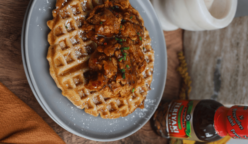A plate with a waffle topped with fried chicken and garnished with herbs, next to a bottle of hot sauce and a marble container on a wooden surface.