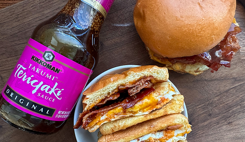 A bottle of Kikkoman Takumi Teriyaki Sauce next to a whole sandwich and a plate with two halves of a sandwich filled with bacon, egg, and cheese on a wooden surface.