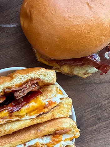 A breakfast sandwich with bacon, egg, cheese, and jam inside a bun, shown whole and cut in half on a wooden surface.
