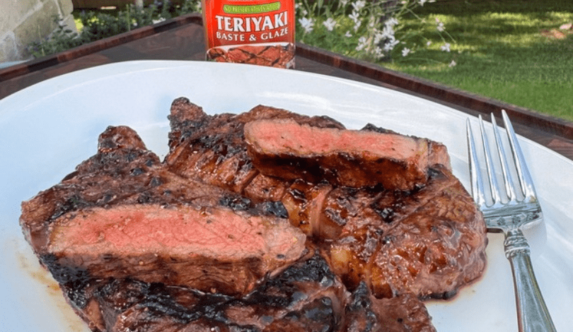Grilled steak slices on a white plate with a fork, accompanied by a bottle of teriyaki baste and glaze in the background.