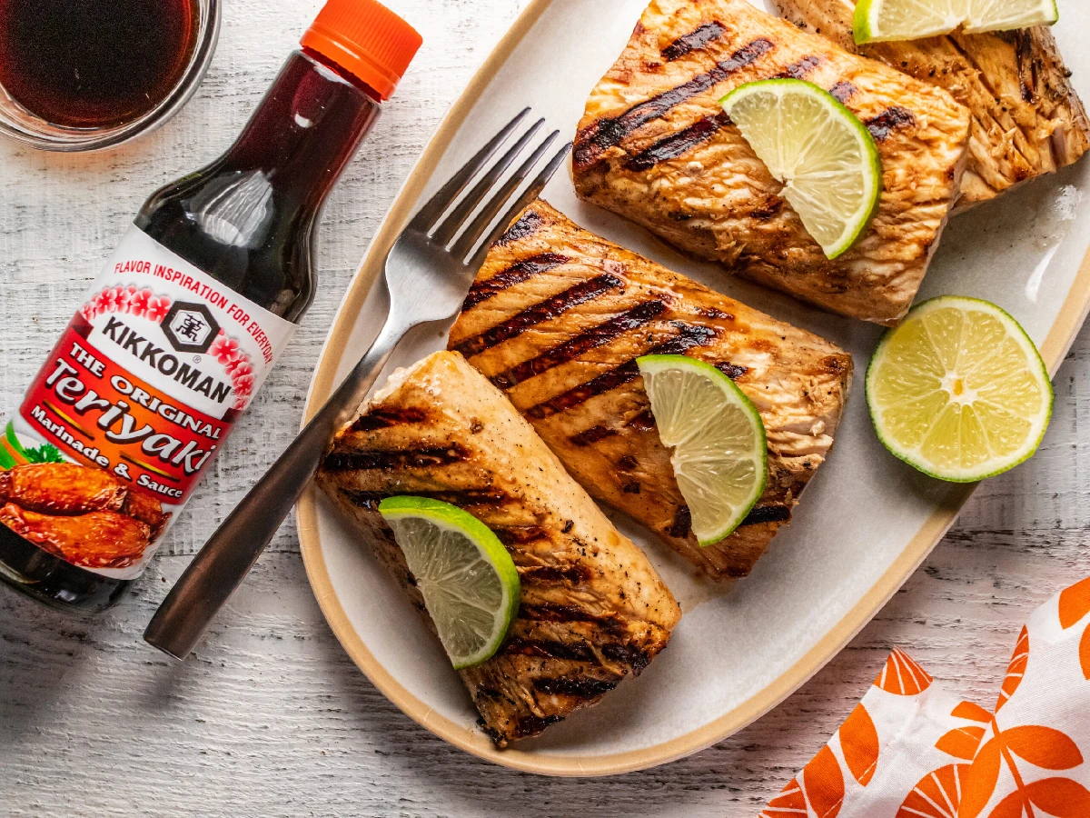 Three grilled fish fillets garnished with lime slices on a plate, with a bottle of Kikkoman Teriyaki marinade and sauce and a glass of dark liquid nearby.