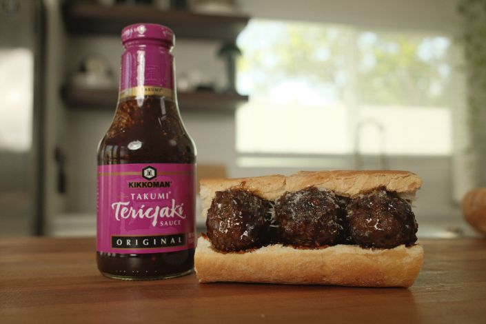 A bottle of Kikkoman Takumi Teriyaki Sauce is next to a sandwich roll filled with three teriyaki-glazed meatballs on a wooden surface.