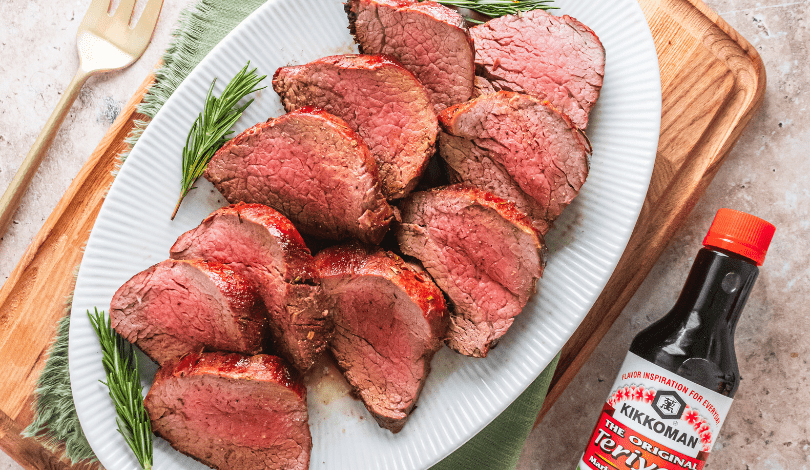 Sliced medium-rare beef arranged on a white platter with rosemary sprigs, next to a bottle of Kikkoman Teriyaki sauce on a wooden board.