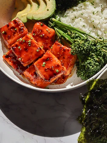A bowl containing rice, broccolini, avocado slices, and glazed tofu cubes, served with a side of seaweed.