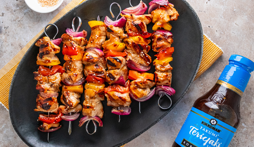 Four grilled chicken and vegetable skewers on a black platter, served with a bottle of Kikkoman Teriyaki sauce and a small dish of sesame seeds.