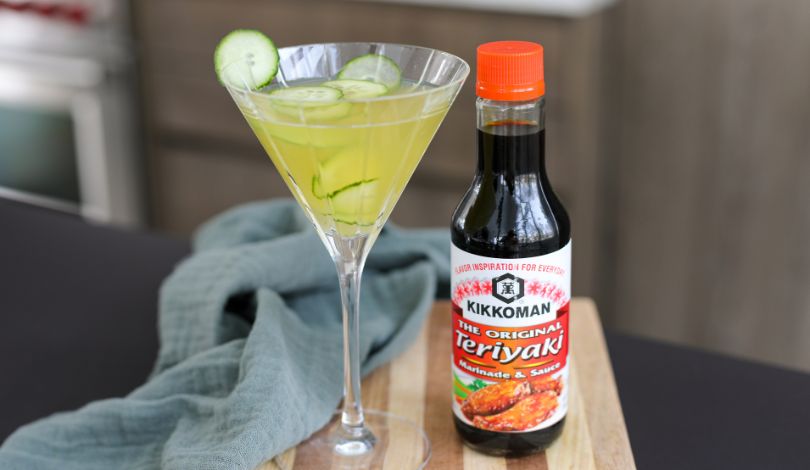 A martini glass filled with a yellow-green drink and cucumber slices sits next to a bottle of Kikkoman Teriyaki Marinade & Sauce on a wooden board.