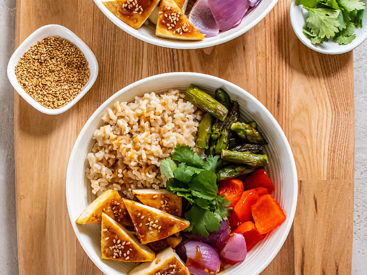 A bowl with brown rice, grilled tofu with sesame seeds, asparagus, carrots, red onion, and cilantro, placed on a wooden surface. A small bowl of sesame seeds and cilantro are nearby.