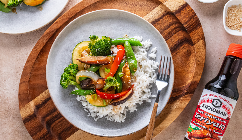 A plate of white rice topped with stir-fried vegetables, next to a bottle of Kikkoman Teriyaki sauce on a wooden board.