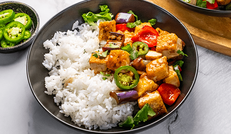 A black bowl with white rice, tofu cubes, eggplant, red bell pepper, jalapeño slices, and cilantro. A small dish of sliced jalapeños is beside the bowl.