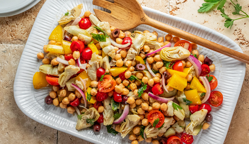 Rectangular platter with a colorful chickpea salad containing cherry tomatoes, yellow bell pepper, red onion, artichoke hearts, olives, and herbs; a wooden serving fork rests on top.