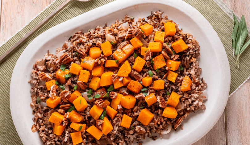 A white platter with a mixture of wild rice, brown rice, roasted butternut squash cubes, and chopped pecans, garnished with herbs.
