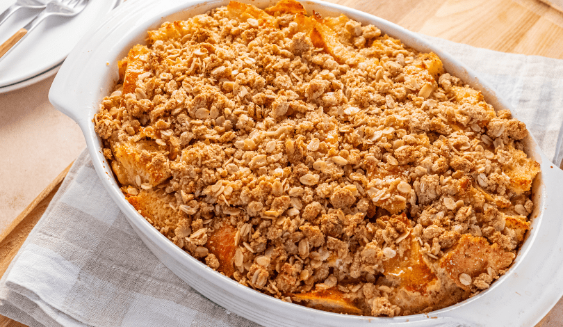 A baked peach crumble in a white oval dish sits on a folded towel atop a wooden surface.