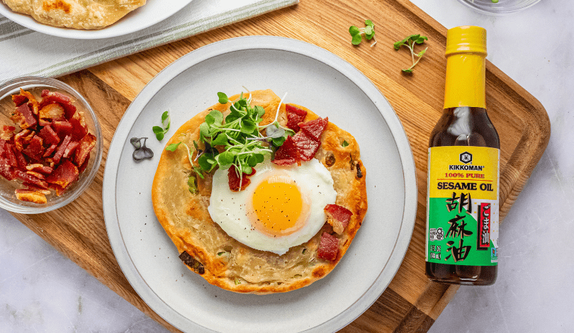A plate with a flatbread topped with a sunny-side-up egg, bacon, and microgreens sits beside a bottle of Kikkoman sesame oil and a small bowl of chopped bacon.