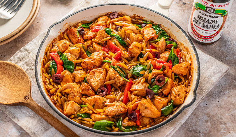 A skillet filled with cooked orzo, chicken pieces, spinach, red bell peppers, and olives, next to a bottle of soy sauce and a wooden spoon on a cloth napkin.