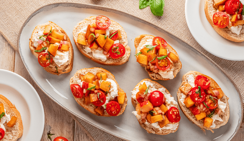 Slices of bread topped with ricotta cheese, diced cantaloupe, cherry tomatoes, and fresh basil are arranged on a gray oval plate and white side plates on a wooden table.