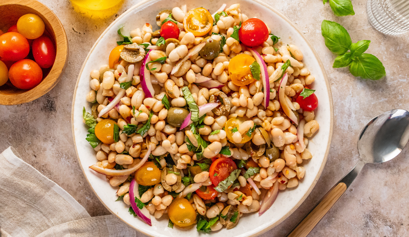 A bowl of white bean salad with cherry tomatoes, red onion, capers, parsley, and olive oil on a light surface, with a spoon and fresh basil nearby.