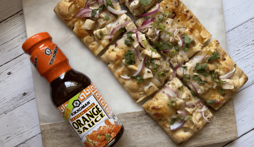 Flatbread topped with chicken, red onion, cilantro, and cheese is sliced and placed next to a bottle of Kikkoman Orange Sauce on a wooden board.