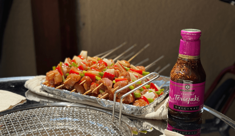 Skewers with marinated meat and vegetables are arranged on foil next to a bottle of Kikkoman Teriyaki sauce and a round grill grate.