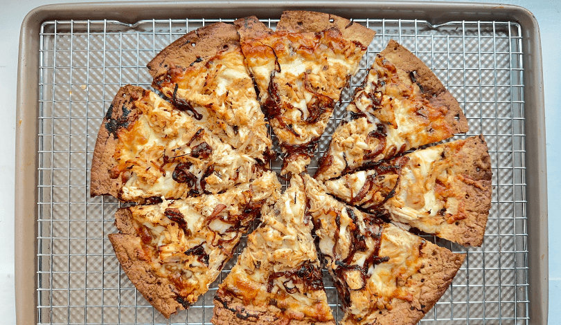 A cooked pizza with barbecue sauce, chicken, and cheese is sliced into twelve pieces on a wire rack over a baking sheet.