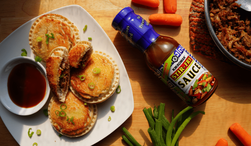 A plate of fried dumplings garnished with green onions, a small bowl of sauce, a bottle of stir-fry sauce, baby carrots, green onions, and a pan of cooked meat on a wooden surface.