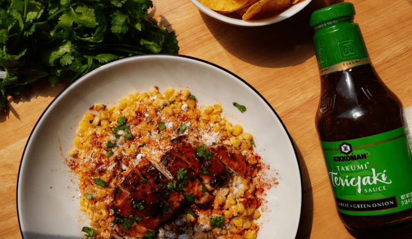 A plate of grilled chicken over corn, sprinkled with herbs and spices, sits beside fresh cilantro, a bowl of tortilla chips, and a bottle of Kikkoman Teriyaki Sauce on a wooden surface.