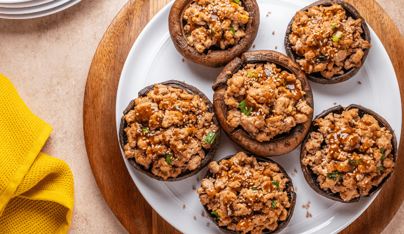 A white plate with six stuffed mushrooms topped with sesame seeds and sauce, placed on a wooden tray next to a yellow cloth.
