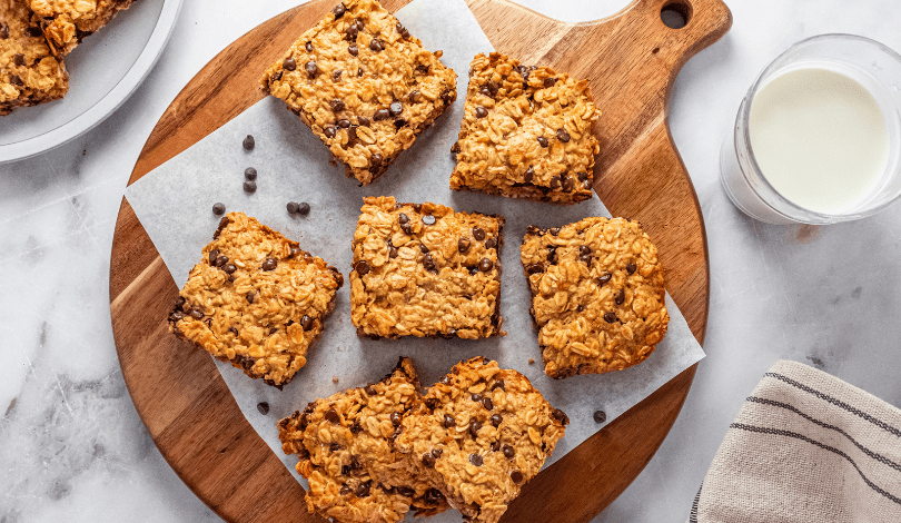 Seven oat and chocolate chip bars are arranged on a round wooden board with parchment paper, next to a glass of milk and a striped cloth napkin.