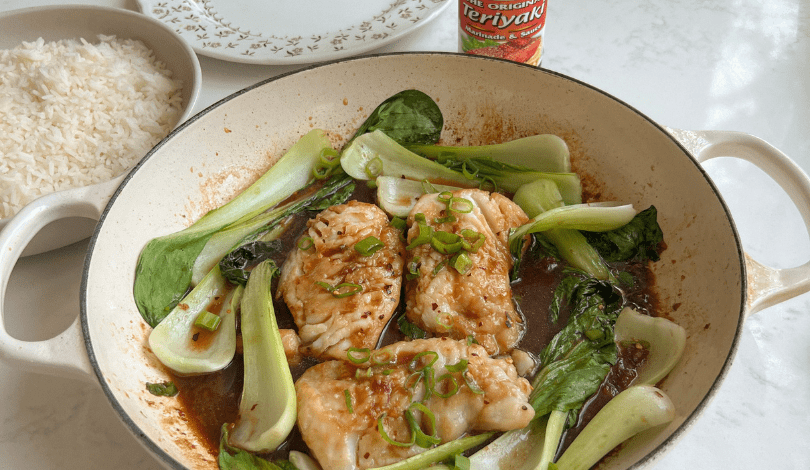 A pan of cooked fish fillets with bok choy in teriyaki sauce, garnished with green onions, sits next to a bowl of white rice and a bottle of teriyaki sauce.