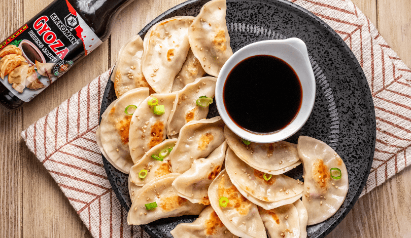 A plate of pan-fried dumplings garnished with green onions and sesame seeds, served with a dish of soy sauce, next to a bottle of gyoza sauce on a striped towel.