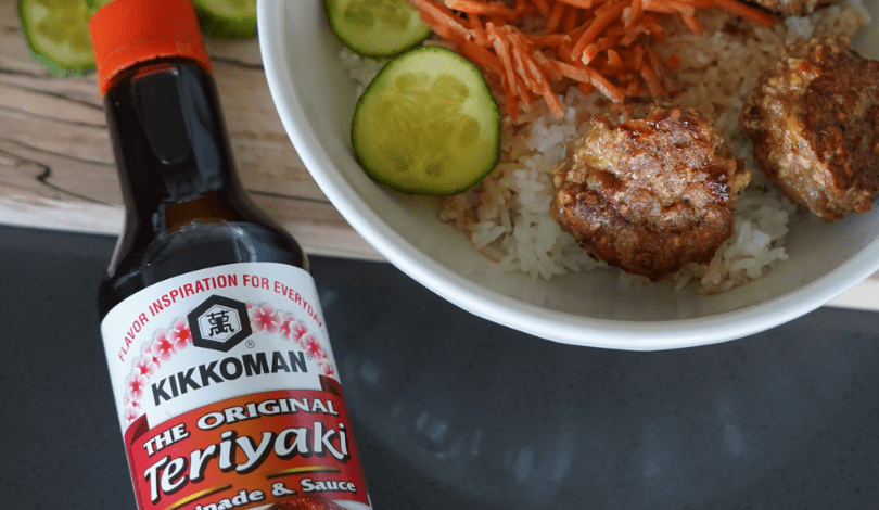 A bowl of rice topped with meatballs, sliced cucumber, and shredded carrots sits next to a bottle of Kikkoman Teriyaki sauce.