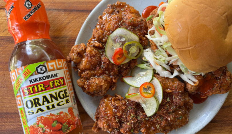 A bottle of Kikkoman Stir-Fry Orange Sauce next to a plate with fried chicken, pickled vegetables, coleslaw, and a sandwich bun.