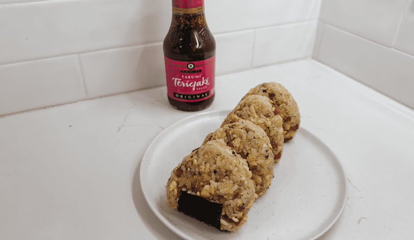 Four rice balls with fillings are arranged on a white plate, with a bottle of Kikkoman Teriyaki sauce in the background on a white countertop.