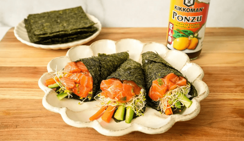 Three salmon and vegetable hand rolls wrapped in seaweed are arranged on a plate, with a stack of seaweed sheets and a bottle of ponzu sauce in the background.