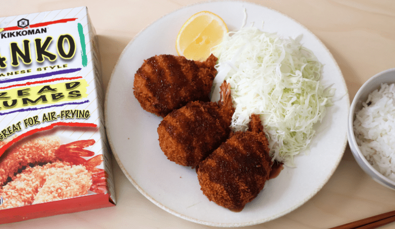 A plate with three breaded shrimp, shredded cabbage, a lemon wedge, and a bowl of white rice next to a box of panko bread crumbs.