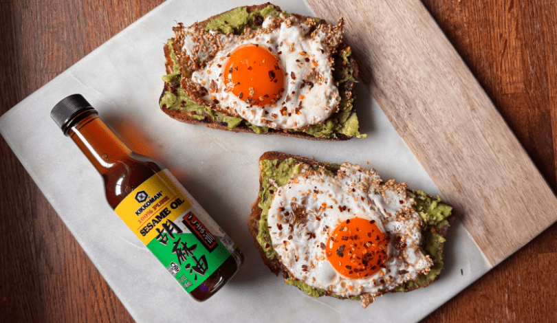Two slices of toast with avocado and sunny-side-up eggs sprinkled with chili flakes, next to a bottle of Kikkoman sesame oil on a marble cutting board.