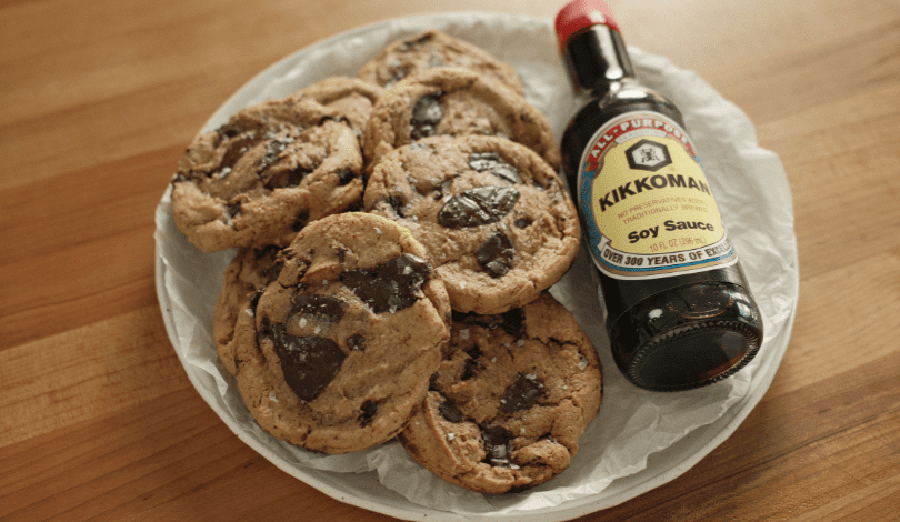 A plate of chocolate chip cookies sits next to a bottle of Kikkoman soy sauce on a wooden surface.