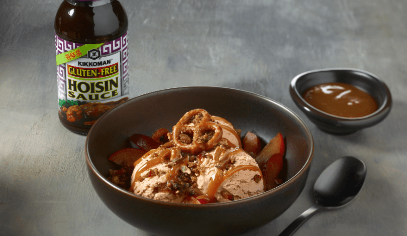 A bowl of ice cream topped with onion rings, chopped nuts, and sauce, with sliced plums; a bottle of gluten-free hoisin sauce and a dish of sauce are beside it.