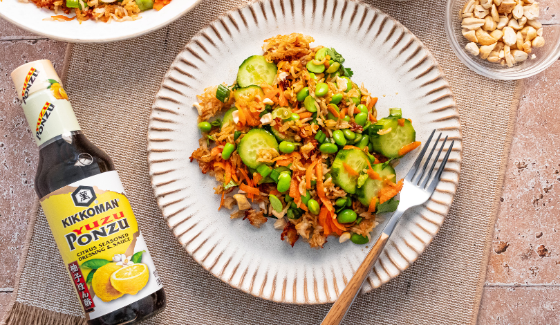 A plate of rice stir-fry with cucumber, edamame, and carrots next to a bottle of Kikkoman ponzu sauce, a fork, and a bowl of peanuts.